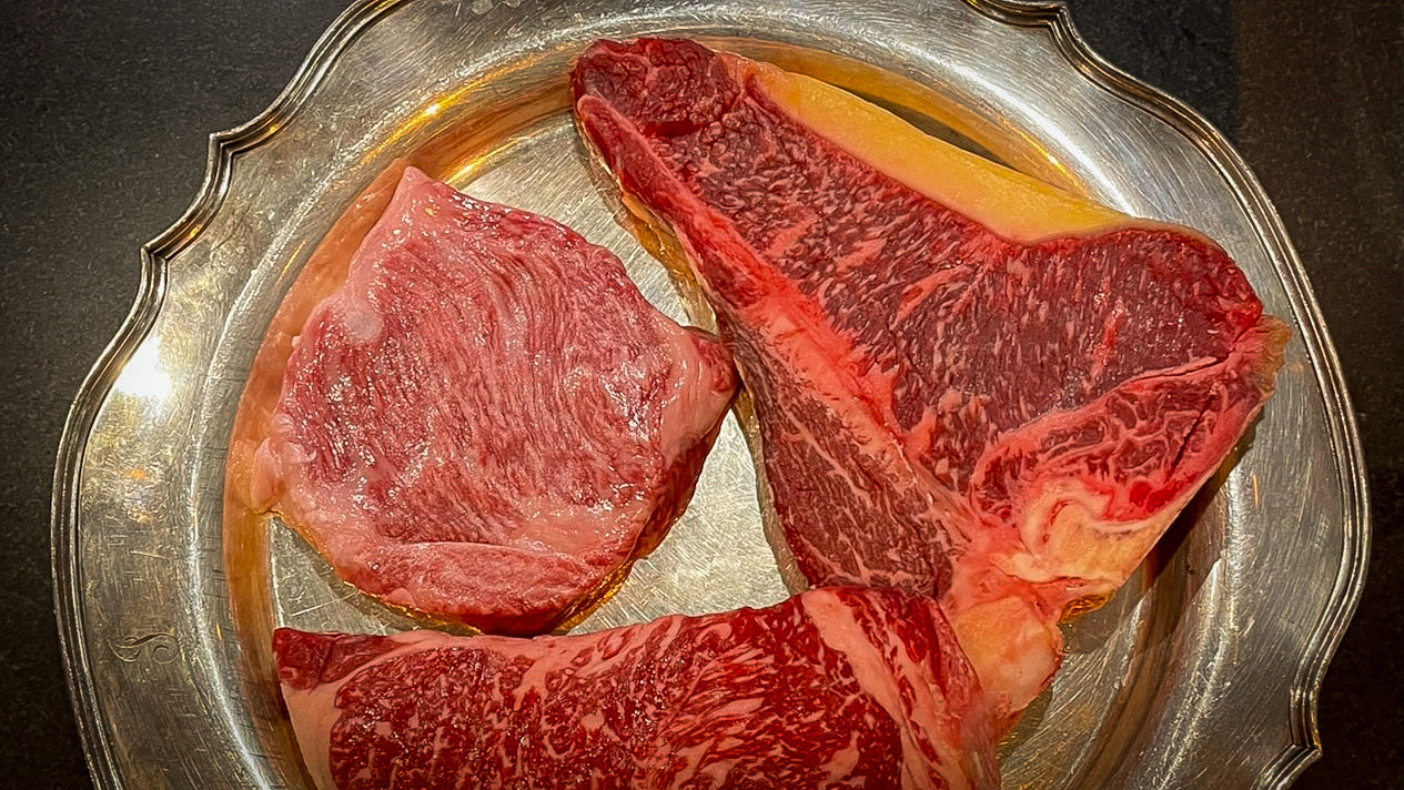 Three pieces of raw steak showing dry aging on a metallic plate with a dark background